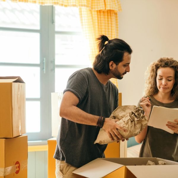 Cheerful young man and woman smiling while unpacking carton boxes with belongings in new apartment during relocation and looking at paper